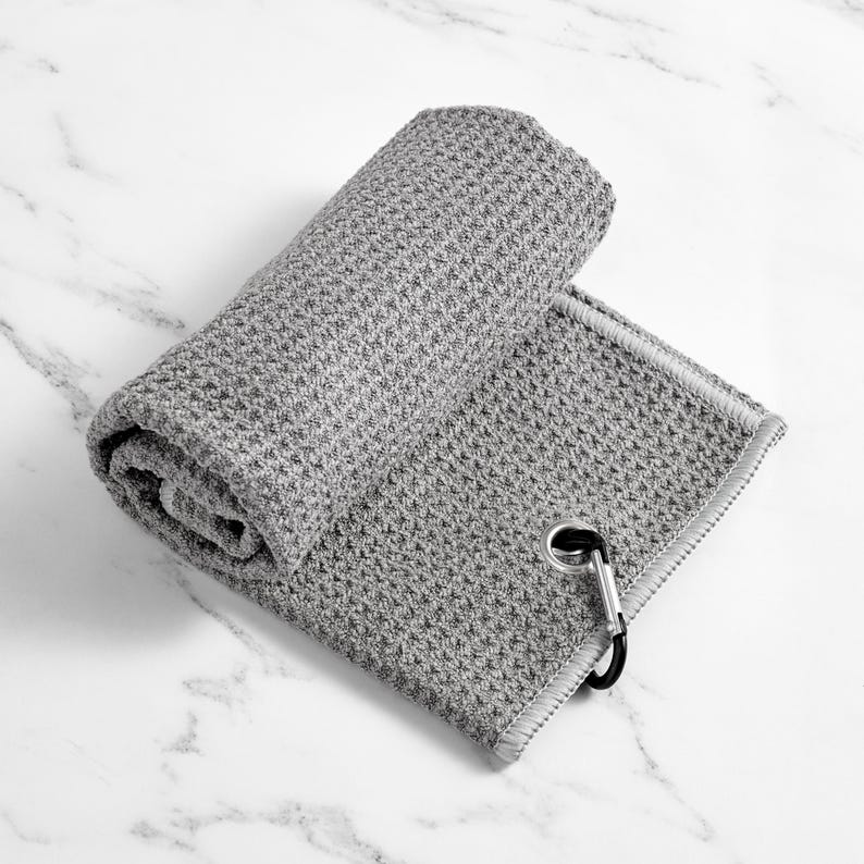May include: A rolled-up grey golf towel with a textured waffle weave. The towel has a silver-coloured grommet and a black carabiner clip for easy attachment to a golf bag. The towel is folded over on a white marble surface.