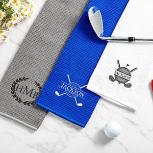 May include: Golf towels in grey, blue, white, and red with custom designs. The grey towel has a monogram, the blue towel says "Jackson", and the white towel features a golf ball and clubs. A golf club rests on the towels.