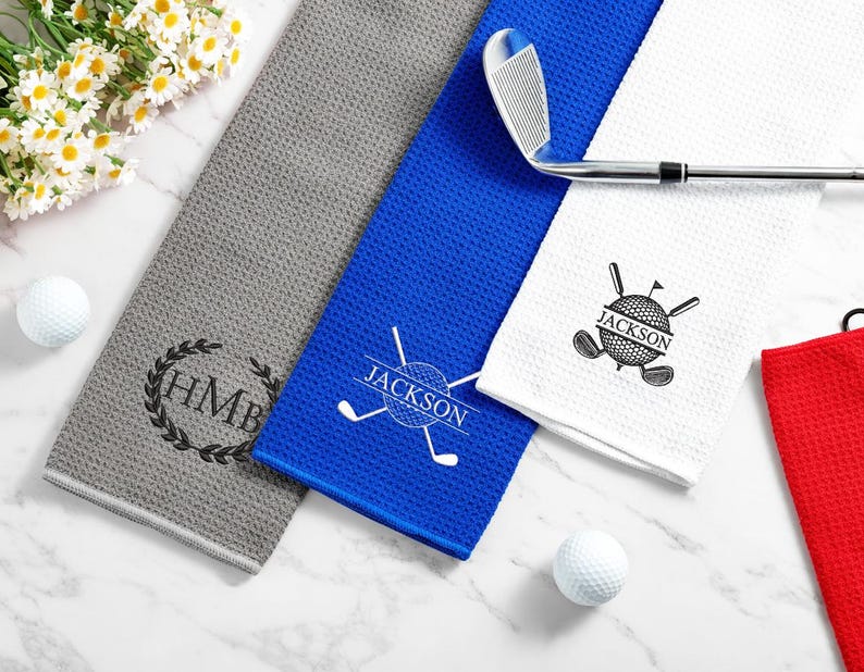 May include: Golf towels in grey, blue, white, and red with custom designs. The grey towel has a monogram, the blue towel says "Jackson", and the white towel features a golf ball and clubs. A golf club rests on the towels.