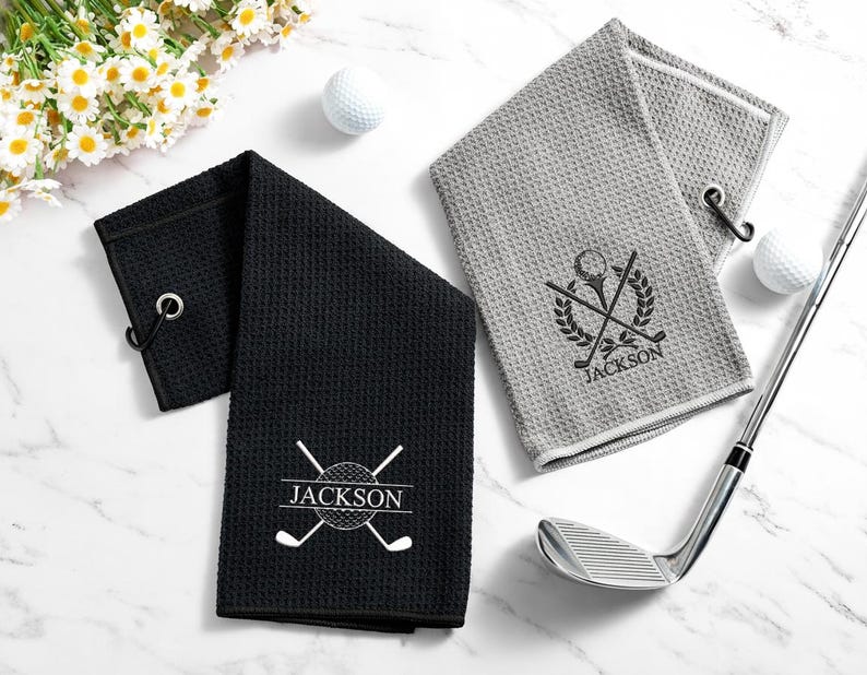 May include: Two golf towels, one black and one grey, with a waffle weave texture. Each towel features a golf-themed emblem with the name "Jackson" and a metal eyelet for hanging. A golf club and golf balls are also present.