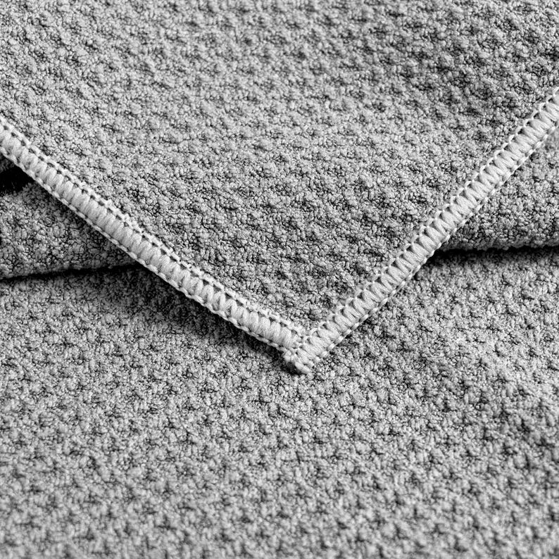 May include: Close-up of a grey, textured fabric with a woven pattern. The edge of the fabric is visible, featuring a white, stitched border. The fabric appears soft and is likely a towel or blanket.