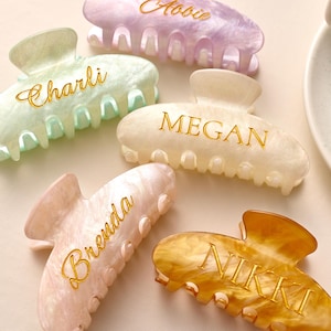 May include: Five personalised hair claw clips in pastel shades: green, lavender, peach, cream, and gold. Each clip features a name in gold script: Charli, Abbie, Brenda, Megan, and Nikki. The clips are made of a marbled, translucent material.