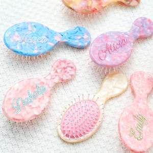 Pode incluir: Uma coleção de escovas de cabelo personalizadas em várias cores pastel. Cada escova apresenta um nome único escrito em letras decorativas. As escovas têm uma pega arredondada e uma almofada de cerdas. Nomes são 'Franca' e 'Alicia'.