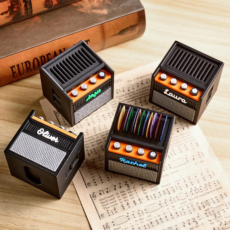 May include: Four miniature black and orange guitar amplifier-shaped boxes. Each box has a speaker grill, knobs, and a name: "Angus", "Laura", "Oliver", and "Rachel". The boxes are on a sheet of musical notes.