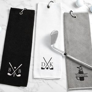 May include: Three golf towels in black, white, and grey, each with a silver carabiner clip. The black towel has the initials "B B" with crossed golf clubs. The white towel has the initials "D K" with crossed clubs and a golf ball. The grey towel has a golf bag design.