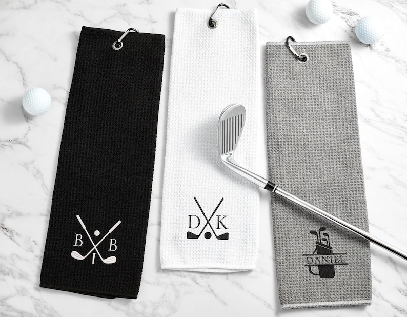 May include: Three golf towels in black, white, and grey, each with a silver carabiner clip. The black towel has the initials "B B" with crossed golf clubs. The white towel has the initials "D K" with crossed clubs and a golf ball. The grey towel has a golf bag design.