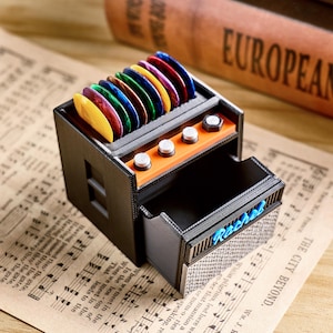 May include: A black and orange guitar pick holder shaped like an amplifier, with a drawer and several colorful guitar picks displayed on top. The word "Rebel" is written in blue on the drawer.