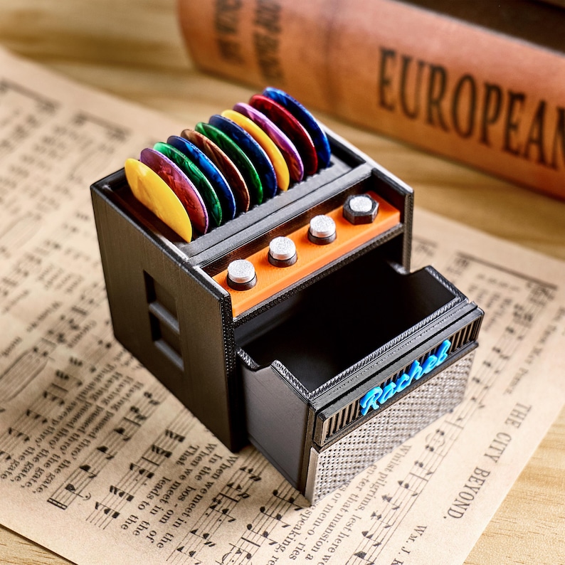 May include: A black and orange guitar pick holder shaped like an amplifier, with a drawer and several colorful guitar picks displayed on top. The word "Rebel" is written in blue on the drawer.