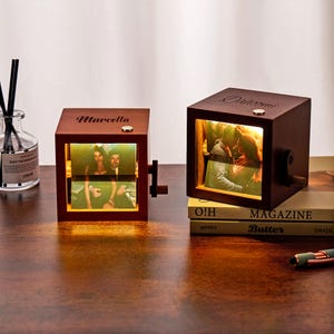 Peut inclure: Deux cubes photo en bois avec des images éclairées. L'un porte l'inscription "Marcella", l'autre "Daheemi". Chaque cube a une manivelle et un petit bouton. Les cubes sont sur une surface en bois.