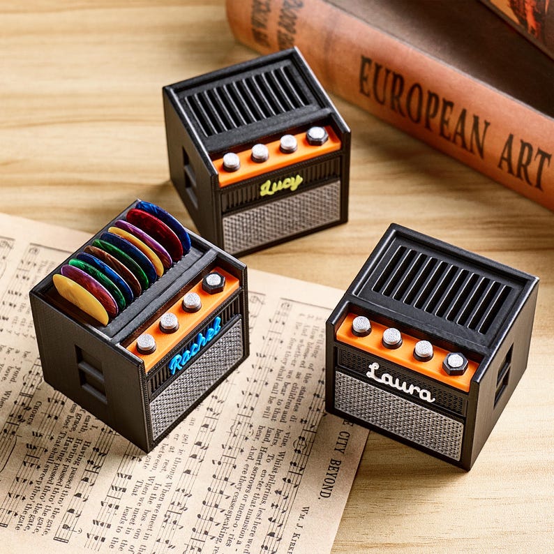 May include: Three miniature black and orange guitar amplifier-shaped boxes. One box holds colorful guitar picks. Each box has a name on it: Lucy, Rachel, and Laura. The boxes sit on sheet music and a wooden surface.
