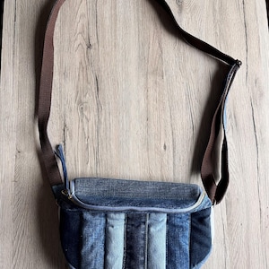Könnte beinhalten: Eine Jeans-Umhängetasche mit braunem Riemen. Die Tasche hat ein Patchwork-Design mit verschiedenen Blautönen. Die Tasche hat einen gebogenen Boden und einen Reißverschluss oben. Der Riemen ist verstellbar und aus braunem Stoff.