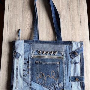Könnte beinhalten: Eine Patchwork-Jeans-Tragetasche in verschiedenen Blautönen. Die Tasche hat eine Reißverschlusstasche, eine Jeans-Tasche mit goldenen Nähten und silberfarbenen Nieten. Die Griffe sind aus Jeansstoff.