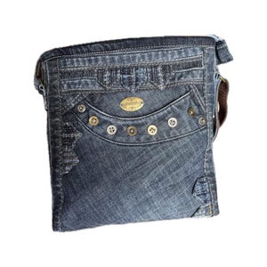 Könnte beinhalten: Eine dunkelblaue Umhängetasche aus Denim in rechteckiger Form mit einer Vordertasche mit geschwungener Klappe und einer Reihe von sechs Metallnieten. Eine ovale, goldfarbene Metallplakette mit eingraviertem Text ist mittig auf der Klappe.