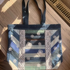 Könnte beinhalten: Jeans-Tragetasche mit Patchwork-Design in verschiedenen Blautönen. Die Tasche hat weiße Spitzenbesätze und kleine weiße Blumenapplikationen. Sie hat zwei Jeans-Tragegriffe und eine Reißverschlusstasche.