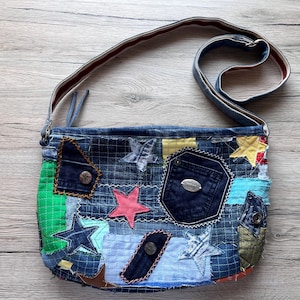 Könnte beinhalten: Eine Patchwork-Umhängetasche aus verschiedenen Blautönen von Jeansstoff. Die Tasche ist mit mehreren applizierten Sternen in Pink, Gelb und Blau sowie einer dunkelblauen Tasche, grünen Stoffakzenten und einem langen, verstellbaren Schulterriemen versehen.