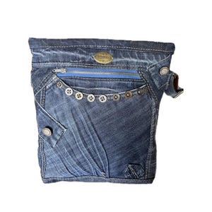 Könnte beinhalten: Eine dunkelblaue Umhängetasche aus Denim mit einer Vordertasche mit leuchtend blauem Reißverschluss, einer Reihe metallischer Knöpfe an der Oberkante, einer kleinen Seitenklappe mit Knopfverschluss und einem ovalen Markenlogo aus Metall oben.