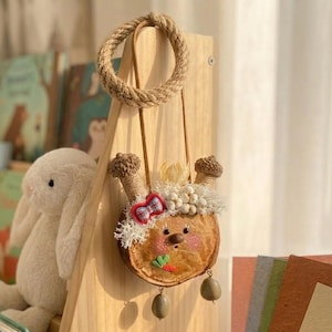 Può includere: Una borsa fatta a mano con un design circolare a fetta di legno. Presenta accenti a forma di ghianda, un fiocco a quadri rosso e blu e perline pendenti verde oliva. La borsa ha un manico in corda ed è decorata con finiture bianche e soffici.