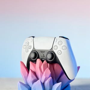 Könnte beinhalten: Ein weiß-schwarzer Gamecontroller ruht auf einem farbenfrohen, kristallförmigen Ständer. Der Ständer wechselt von Blau zu Rosa und erzeugt eine lebendige Darstellung. Der Controller verfügt über Tasten-Symbole und ein zentrales Logo.
