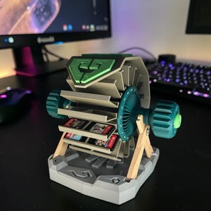 Op de afbeelding: Een grijze en teal 3D-geprinte spelcartridgehouder. De houder heeft een ventilatorachtig ontwerp met een groene bovenkant en teal accenten. De houder is ontworpen om meerdere spelcartridges te bevatten. De basis is grijs en de steunen zijn van hout.