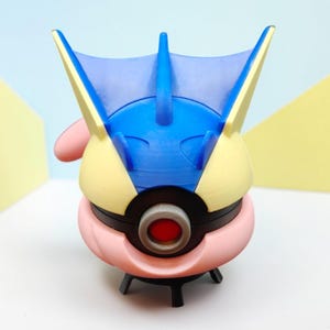 Könnte beinhalten: Eine farbenfrohe, 3D-gedruckte Figur eines Pokémon-Charakters. Die Figur hat einen blauen Kopfkamm, einen gelben und rosa Körper und ein rotes Auge. Sie befindet sich vor einem hellblauen und gelben Hintergrund.