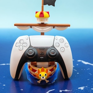 Könnte beinhalten: Ein weiß-schwarzer Videospiel-Controller mit einem Piratenschiff-Design. Das Schiff hat eine schwarze Flagge, ein gelb-rotes Segel und eine Cartoon-Figur als Galionsfigur. Der Controller befindet sich vor einem blauen Hintergrund.