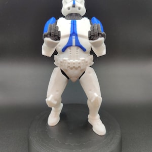 May include: A white and blue action figure of a clone trooper from a science fiction franchise. The figure is standing on a black circular base. The trooper's armor has blue accents on the helmet, shoulders, and chest. The figure is posed with its hands up.