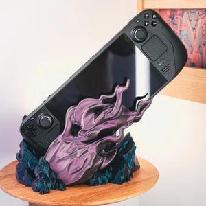 May include: A black handheld gaming console rests on a custom stand. The stand has a purple and teal flame design, placed on a wooden shelf. The shelf is supported by a wooden bracket, with a white wall in the background.