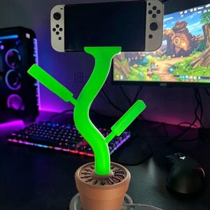 Può includere: Un supporto verde a forma di pianta per telefono o dispositivo di gioco. Il supporto è progettato per assomigliare a una pianta in un vaso marrone, con un dispositivo appoggiato sopra. Si trova su una superficie nera, con una tastiera e un monitor per computer sullo sfondo.