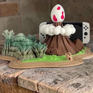 Può includere: Un supporto per console di gioco a forma di vulcano marrone con una parte superiore a forma di uovo bianco e rosso, nuvole bianche e una base verde e beige. Il supporto contiene una console di gioco bianca e nera. La base presenta erba verde, alberi e un piccolo castello di sabbia.