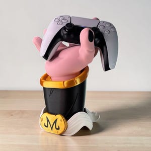 Könnte beinhalten: Ein rosa handförmiger Controller-Ständer, der einen weiß-schwarzen Gamecontroller hält. Der Ständer hat einen schwarzen Sockel mit goldenem Rand und ein gelbes Emblem mit dem Buchstaben "M".