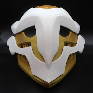 May include: A white and gold 3D printed mask with a detailed design. The mask has a sharp, angular shape with a pointed top and a curved bottom. The mask is made of a smooth, plastic-like material.
