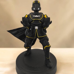 Könnte beinhalten: Eine schwarz-gelbe Soldatenfigur in taktischer Rüstung mit Helm, Umhang und erhobenen Händen. Die Figur trägt ein weißes Totenkopf-Emblem auf Brust und Gürtel und steht in einer dynamischen Pose auf einem runden, schwarzen Sockel.