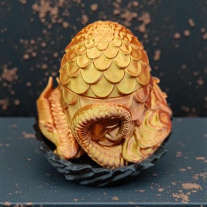 May include: A golden dragon egg sculpture with a textured, scaled surface. The egg rests in a dark, nest-like base. The sculpture has a detailed, ornate design and is set against a dark background.