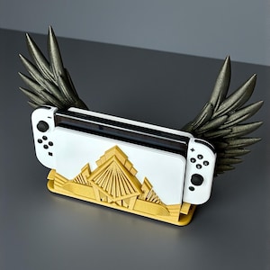 Può includere: Console per videogiochi bianca e dorata con pulsanti neri, appoggiata su un supporto dorato con un design a piramide. La console è affiancata da ali grigie metallizzate, creando un'estetica unica e decorativa.