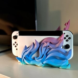 Può includere: Una console Nintendo Switch bianca inserita in un supporto personalizzato con un design a fiamme scolpite in tre dimensioni. L'accessorio a forma di fiamma sfuma dal colore azzurro alla base fino a un viola vivace nella parte superiore.