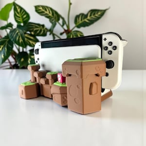 May include: A white and black Nintendo Switch console is displayed on a brown and green landscape-themed stand. The stand features a pixelated design with a red block. The console is positioned upright.