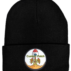 May include: Black beanie with a circular patch featuring a rocket ship design. The patch includes the text "Custom Patch" and space for a logo or image. The beanie is made of a knitted material.