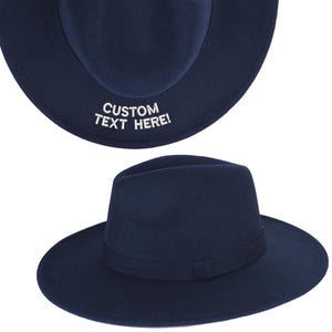 May include: A navy blue fedora hat with a thin black band around the crown. The hat has the text "CUSTOM TEXT HERE!" embroidered on the brim.