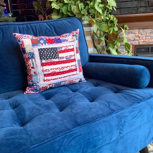 May include: A square pillow with a patchwork design featuring the American flag. The pillow has a white border and is placed on a blue velvet couch. The flag design includes red and white stripes and a field of blue with white stars.