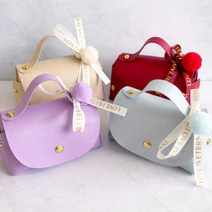 Pode incluir: Quatro pequenas bolsas em tons pastel com alças e ferragens douradas. Cada bolsa é adornada com uma fita que diz "LOVE IS ETERN" e um pompom a condizer. As bolsas são em tons de lavanda, bege, vermelho e azul claro.