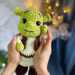 Puede incluir: Muñeco de ganchillo hecho a mano del personaje Shrek. El muñeco es verde con una camisa blanca, un chaleco marrón y zapatos marrones. El muñeco tiene ojos negros y una sonrisa cosida. El muñeco está sostenido por dos manos.