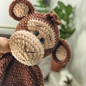 May include: A handmade plush monkey in shades of brown and tan. The monkey has a textured, fuzzy appearance, with a light tan face and ears, and dark brown accents. The eyes are blue, and the mouth is stitched with black thread. A soft toy.