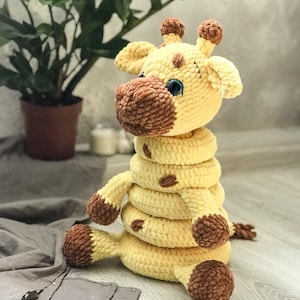 May include: A crocheted giraffe toy in yellow and brown. The giraffe features a segmented neck, brown spots, and blue eyes. The toy is designed to sit upright. The toy is on a light-colored surface.