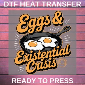 May include: DTF heat transfer design featuring the text "Eggs & Existential Crisis" in orange and black. The design includes a skillet with two fried eggs. The words "Ready to Press" are at the bottom.