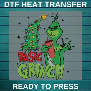 May include: A green Grinch character holding a red cup and a candy cane next to a decorated Christmas tree. The words "BASIC GRINCH" and "READY TO PRESS" are in the image. DTF heat transfer.