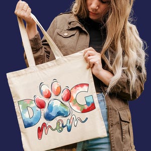 May include: A white canvas tote bag with a colorful watercolor design of a paw print and the words "Dog Mom".