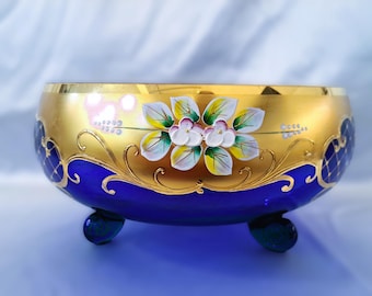 May include: A cobalt blue glass bowl with gold trim and floral accents. The bowl has three feet and is decorated with a gold and white floral design.