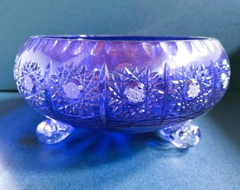 May include: A deep blue, cut-glass bowl with a rounded shape and three small feet. The bowl features intricate starburst and floral patterns carved into the glass. The bowl is a decorative item, likely for display or serving.
