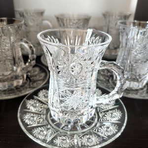 Può includere: Un set composto da tazza da tè e piattino in vetro trasparente con intricati motivi a stella e tagli geometrici. La tazza presenta un manico curvo e un bordo svasato, mentre il piattino abbinato mostra un motivo a stella radiale ripetuto.