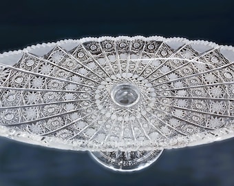 May include: A clear glass serving dish with a pedestal base. The dish has a scalloped edge and a delicate, intricate pattern.